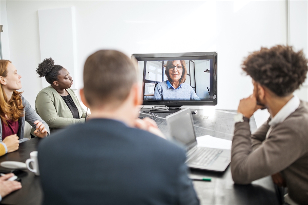 Picture of a group of people sitting at a conference table and looking at a screen with a person video calling in