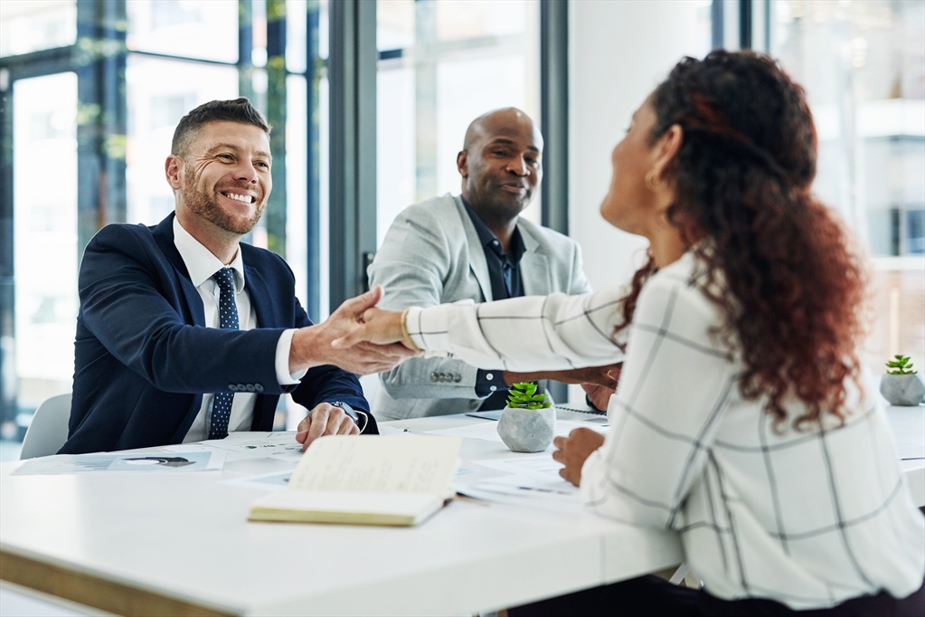 Picture of two people sitting on the same side of the table while one smiles and shakes hands with a person on the other side